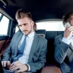 Two corporate executives working on laptop and phone in backseat of luxury chauffeur-driven car, representing premium business travel services.