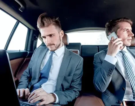 Two corporate executives working on laptop and phone in backseat of luxury chauffeur-driven car, representing premium business travel services.