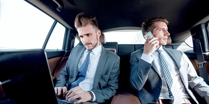 Two corporate executives working on laptop and phone in backseat of luxury chauffeur-driven car, representing premium business travel services.