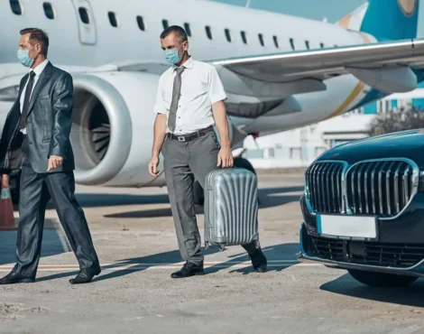 Business travelers in suits wearing masks walking from airplane toward luxury chauffeur car, highlighting safe and professional airport transfer service.
