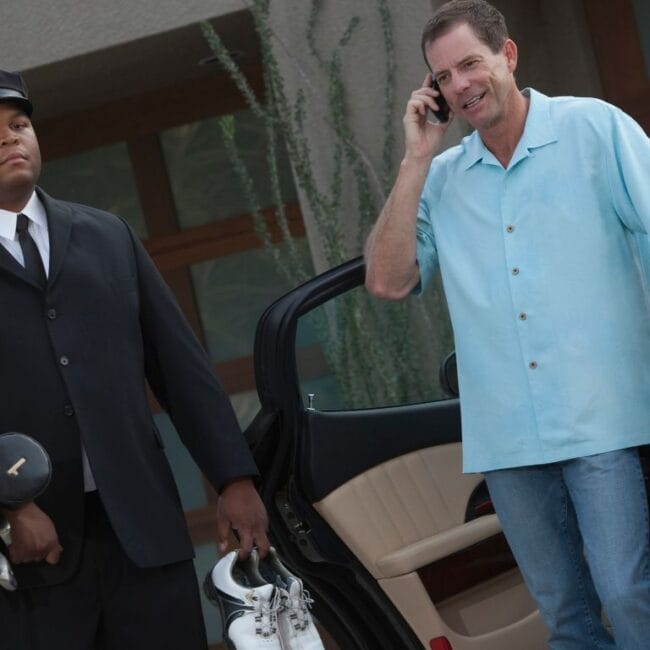 A uniformed chauffeur carrying golf clubs and shoes next to a man on a cell phone by an open luxury car door.
