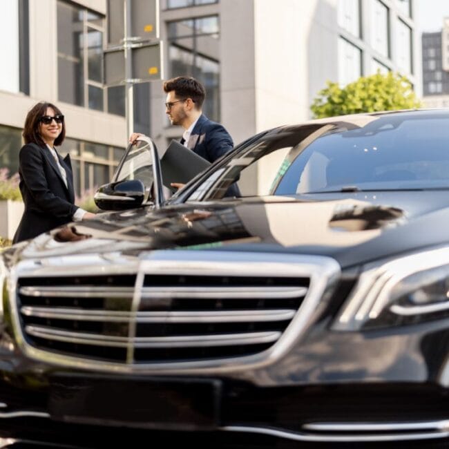 Close-up of a black luxury car with a chauffeur in a suit holding the door open for a professional woman in sunglasses.