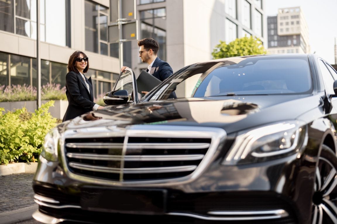 Close-up of a black luxury car with a chauffeur in a suit holding the door open for a professional woman in sunglasses.