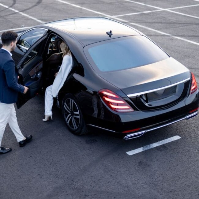 High-angle view of a chauffeur in a blazer holding the door of a black luxury sedan for a female executive in a white suit in a parking lot.