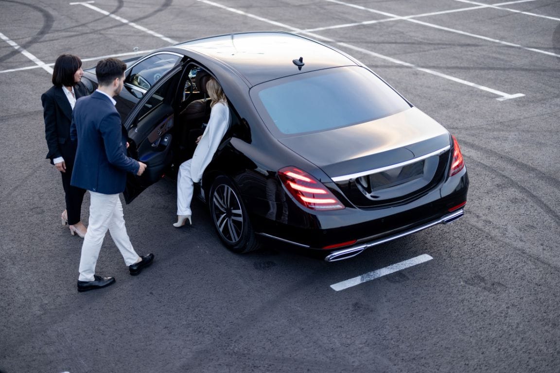 High-angle view of a chauffeur in a blazer holding the door of a black luxury sedan for a female executive in a white suit in a parking lot.