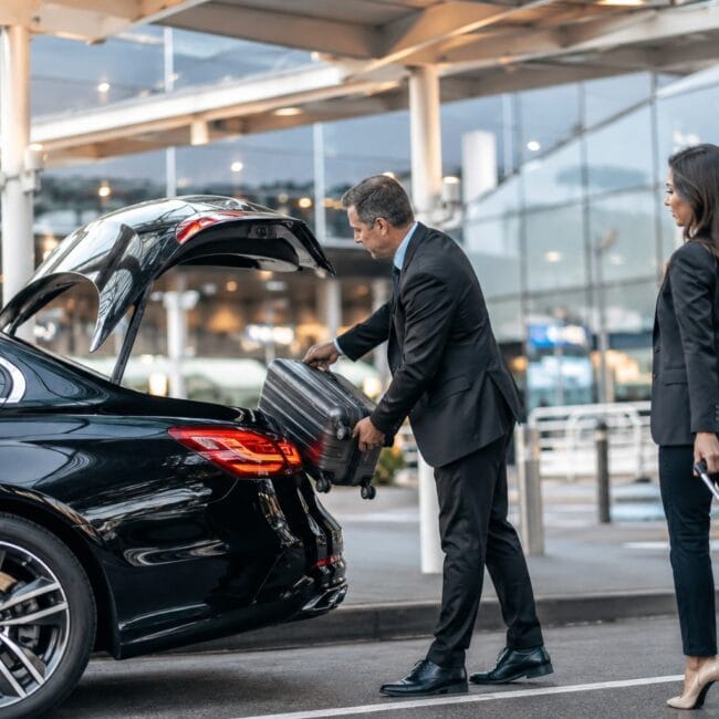 a professional chauffeur in a black suit loads a suitcase into the trunk of a luxury black car outside an airport terminal while a businesswoman stands nearby with her rolling luggage.