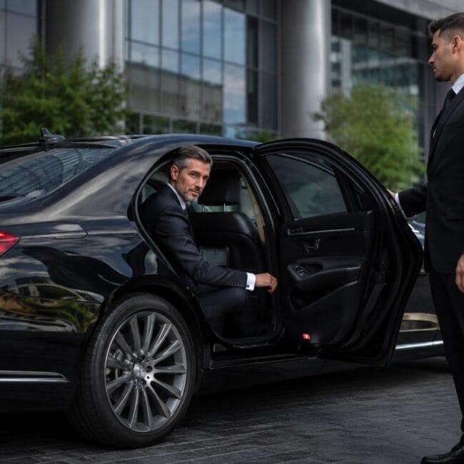 An executive in a suit looks out from the backseat of a luxury black sedan while a chauffeur in a black suit holds the door open outside a modern office building.